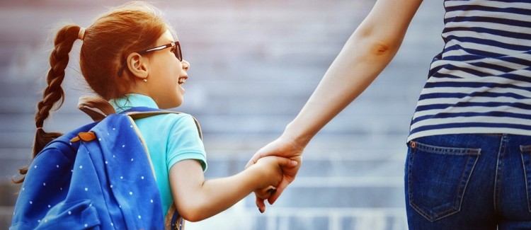 young girl in glasses wearing blue backpack holds hand of adult woman as she goes back to school